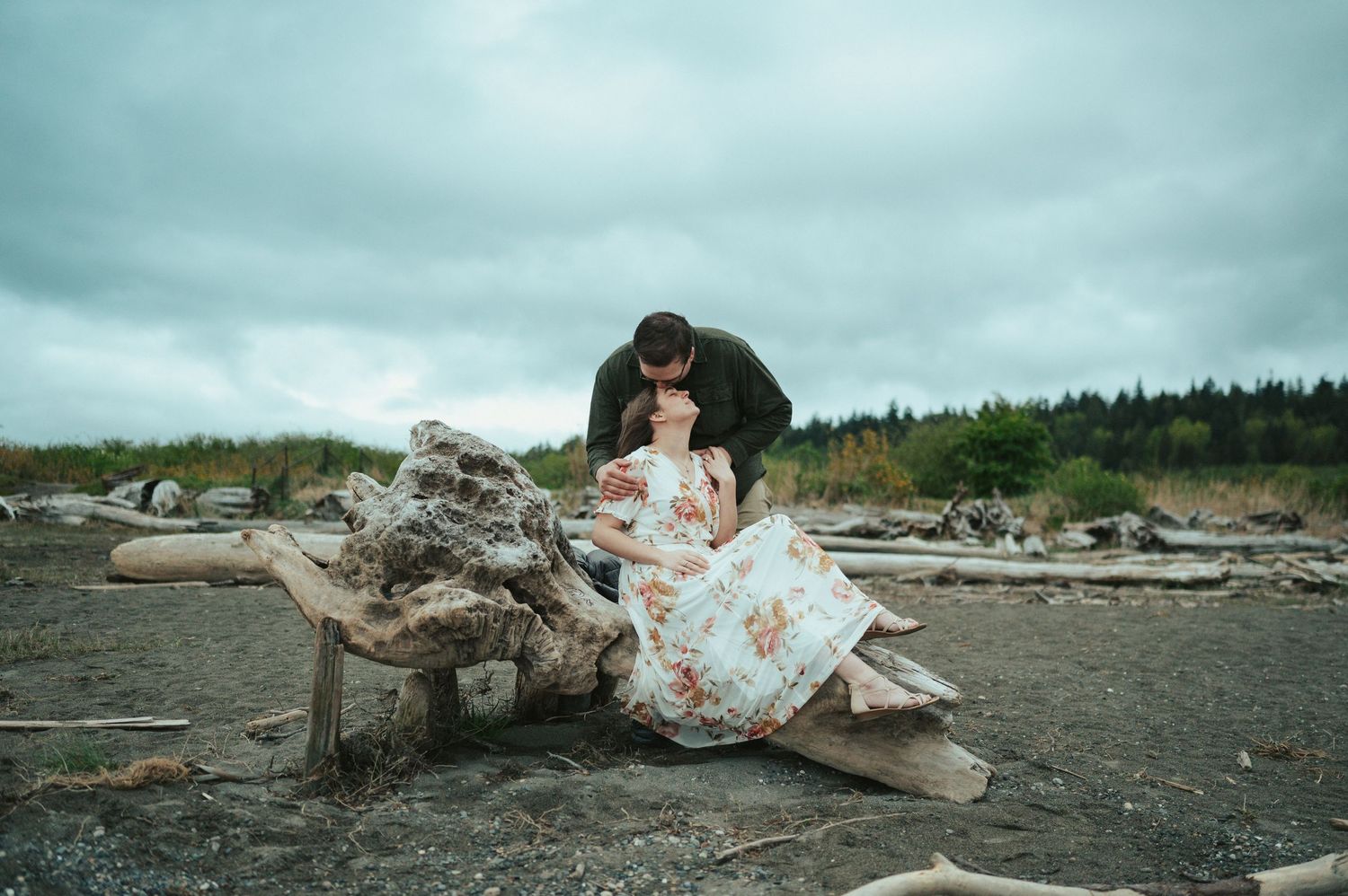 A romantic couple shares an intimate moment while sitting on a large piece of driftwood at the beach.