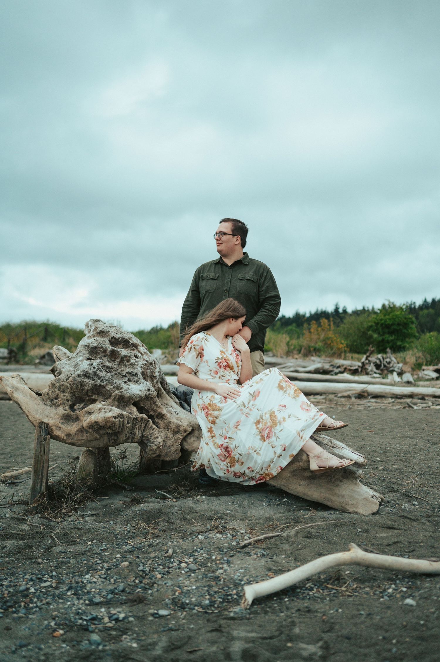 A couple sits on driftwood at a moody beach with cloudy skies and scattered logs in the background.