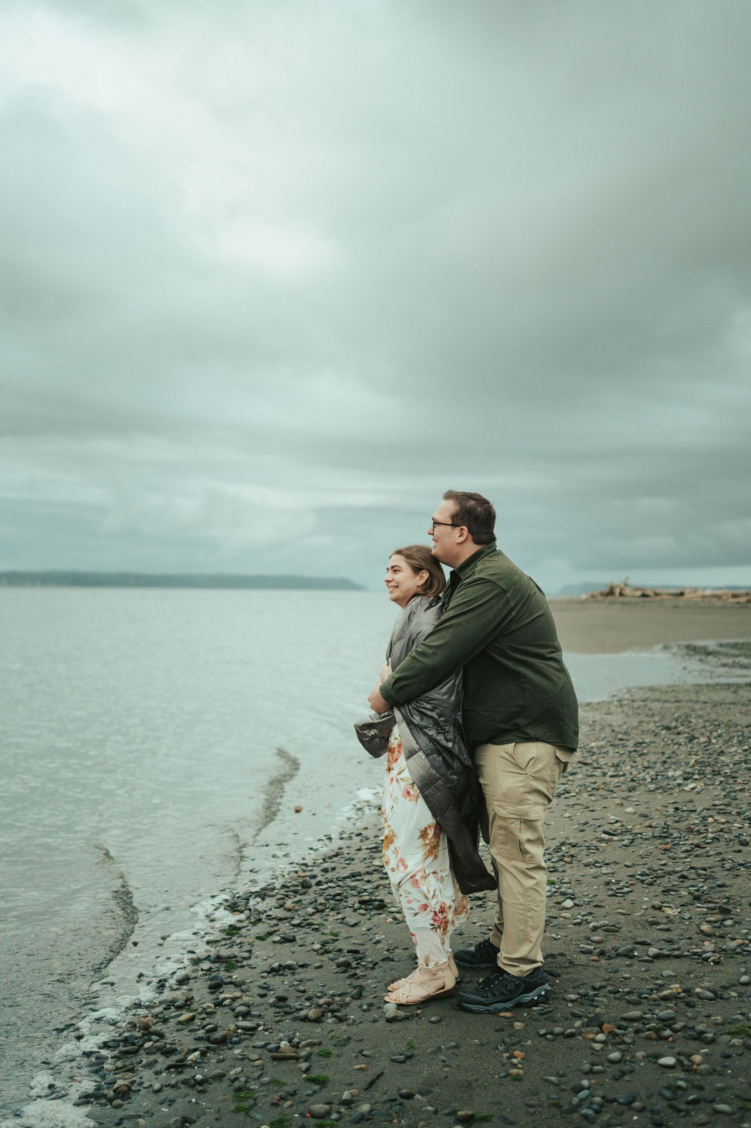 A couple embraces on a rocky beach shore against a moody grey sky and calm waters.