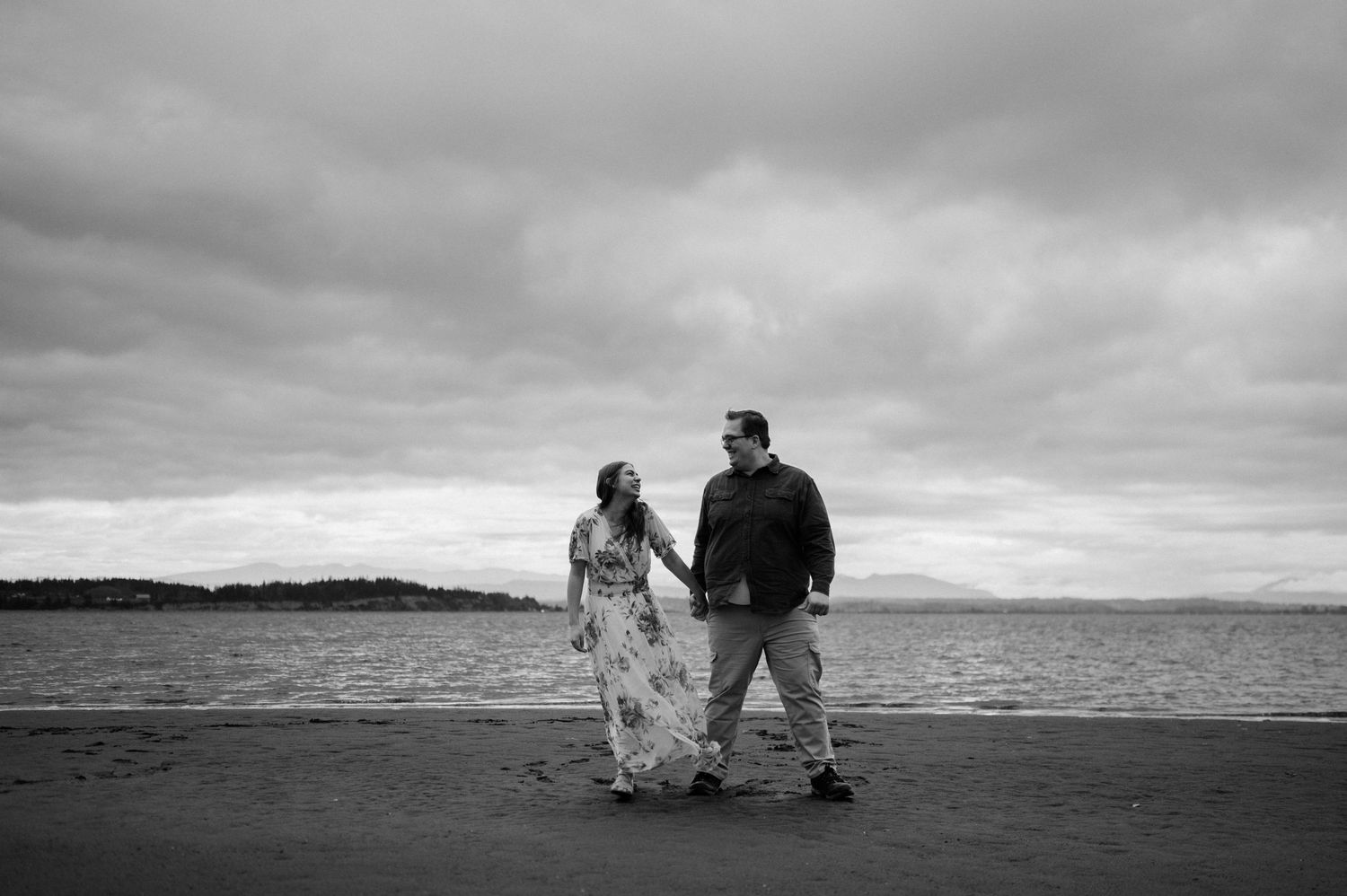 A couple stands together on a moody beach shoreline with dramatic cloudy skies in black and white.