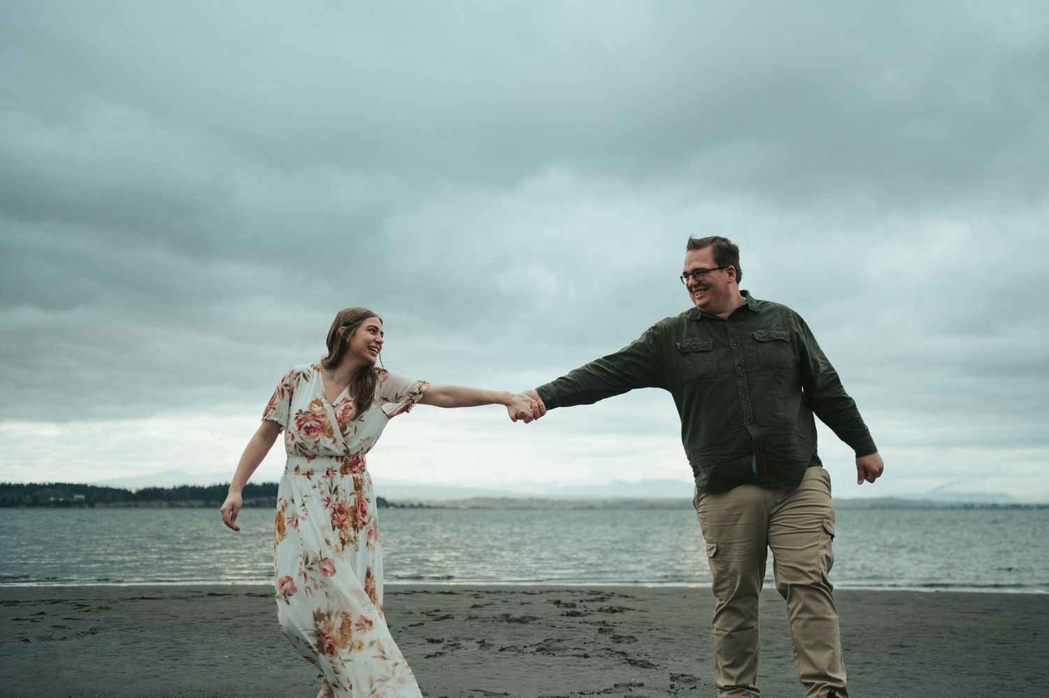 A couple holds hands while walking along a misty beach with dramatic overcast skies.