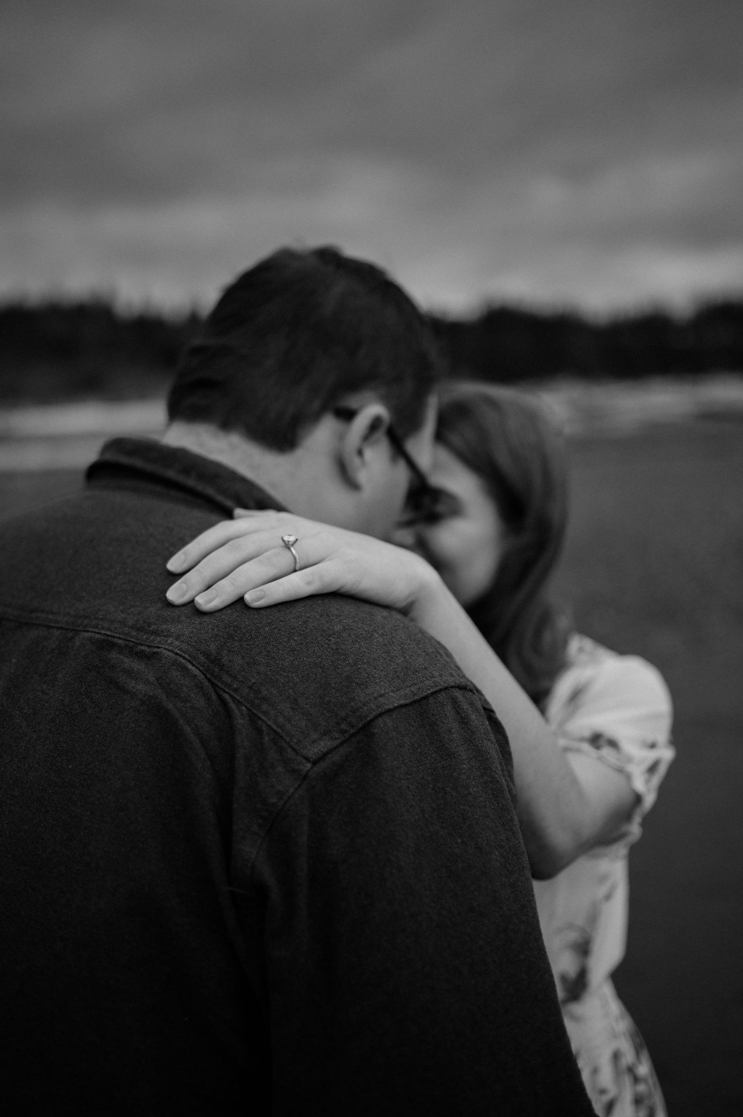 A romantic black and white photo showing an intimate embrace on a cloudy beach, with a wedding ring visible.