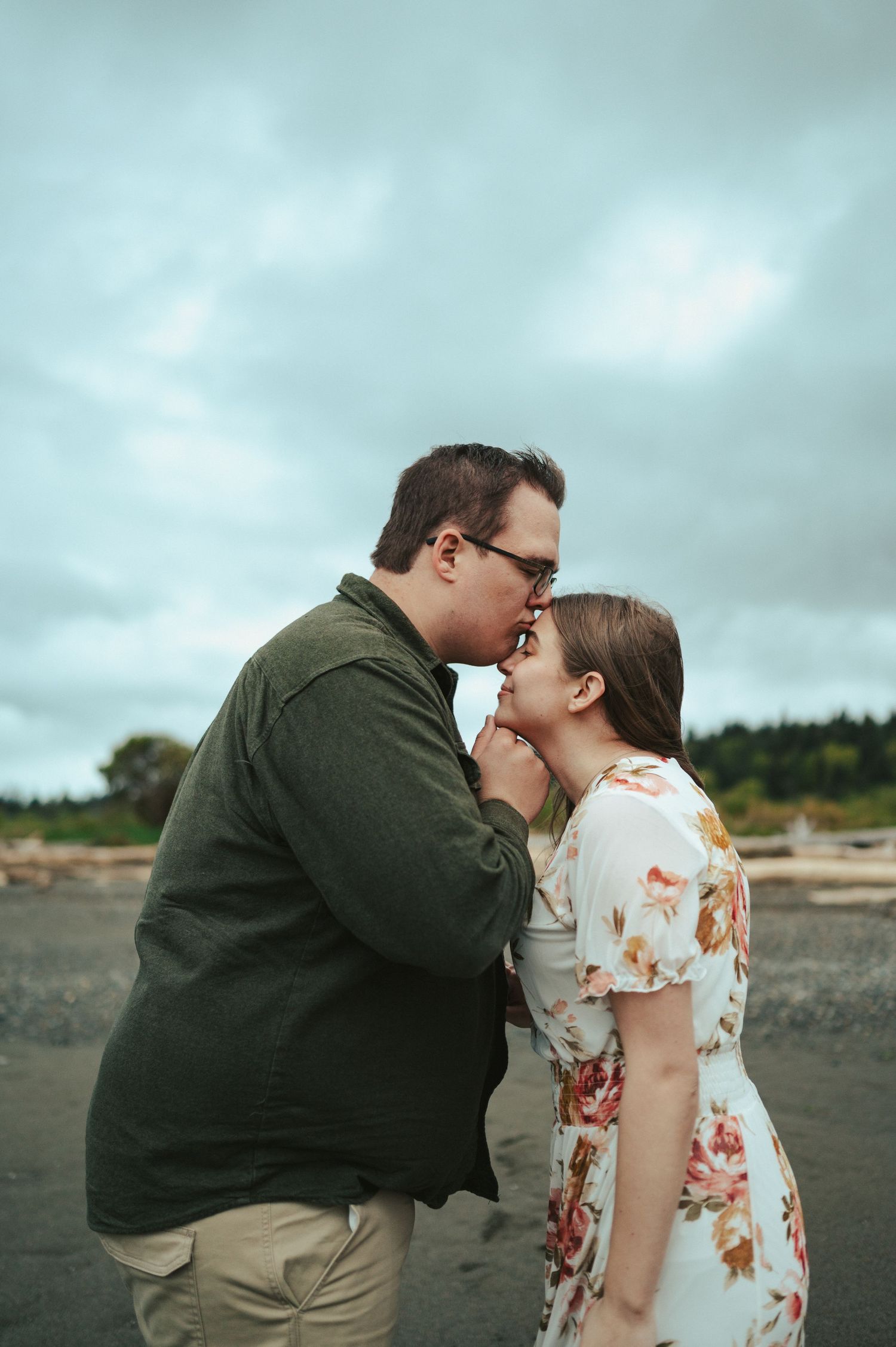 An intimate couple shares a tender moment on a cloudy beach while wearing casual clothing.