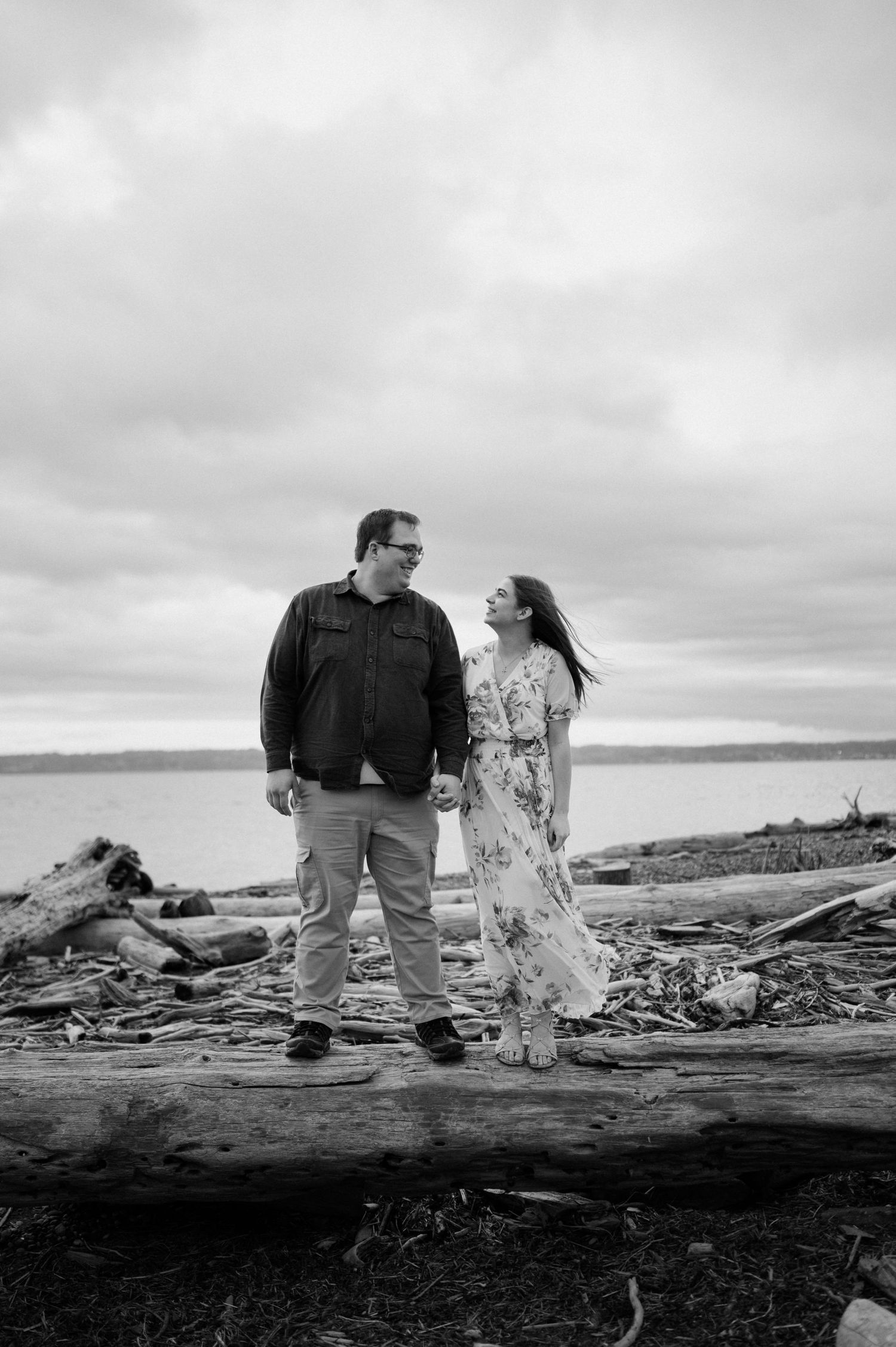 A couple stands together on driftwood logs at a cloudy beach with water in the background.