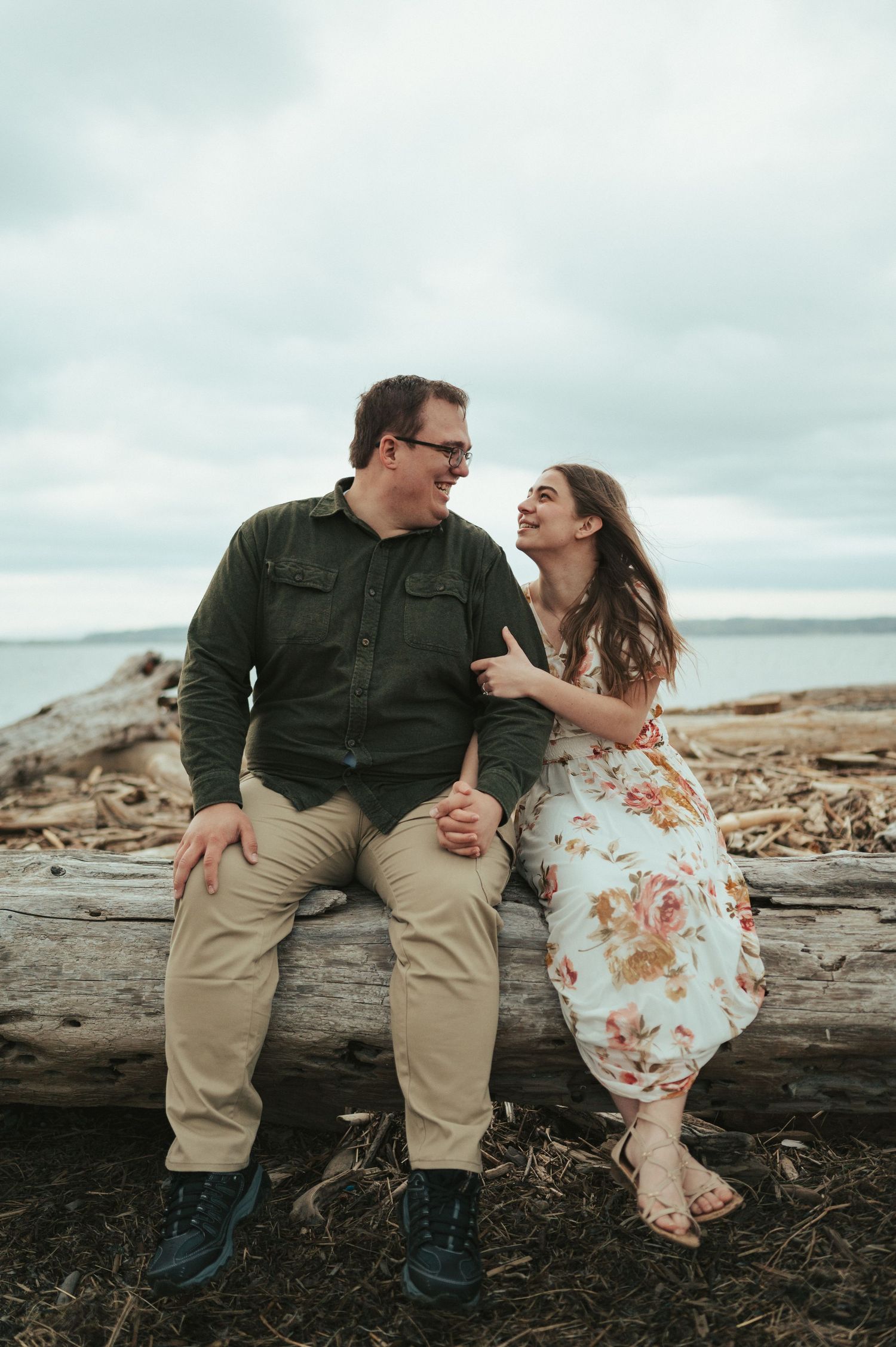 A couple shares a playful moment while sitting on driftwood at a rocky beach under overcast skies.