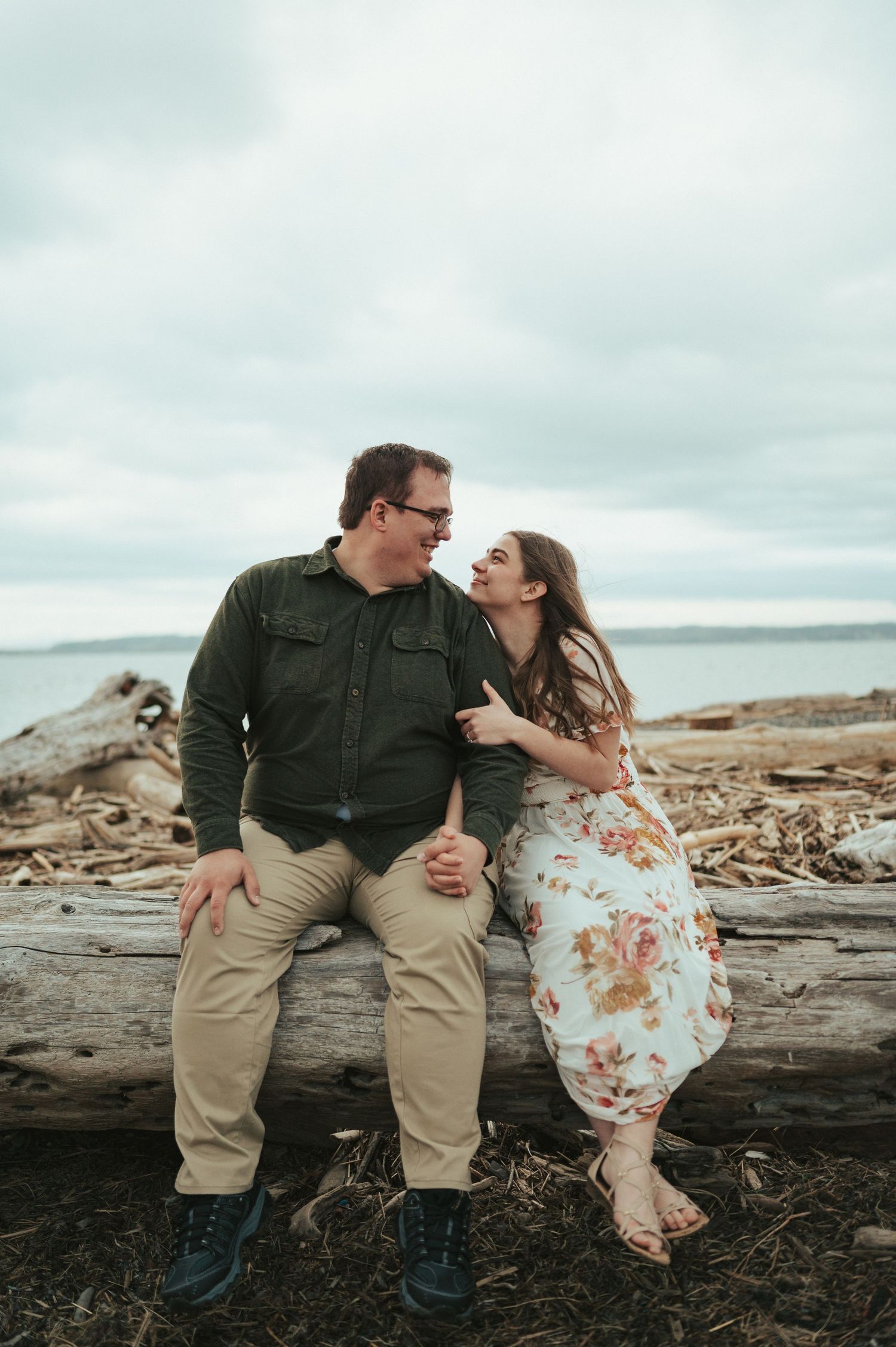 Couple sharing a romantic moment while sitting on driftwood at a moody beach under overcast skies.