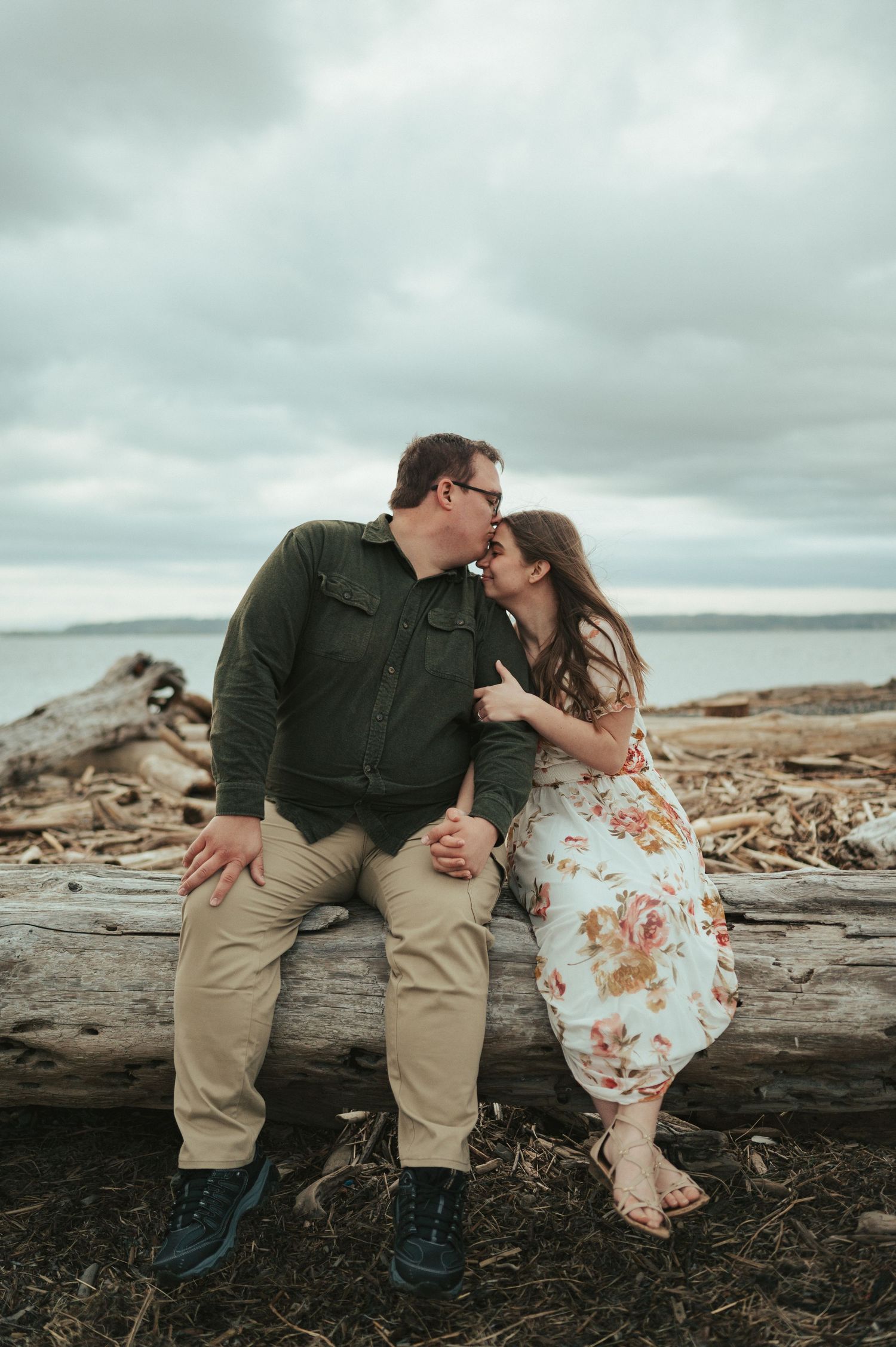 A romantic couple shares an intimate moment while sitting on driftwood at a cloudy beach.