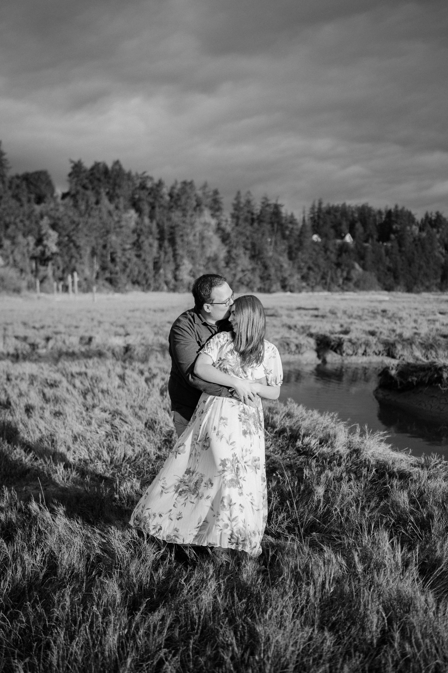 A romantic black and white portrait of a couple embracing in a meadow with forest and river views.