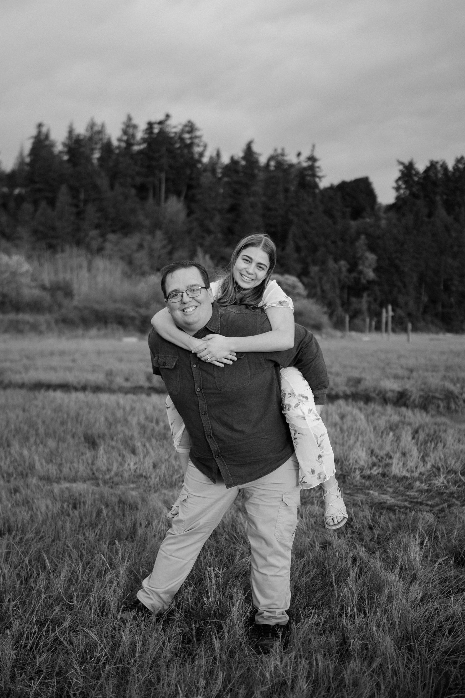 A playful black and white portrait of a couple posing together in a field with evergreen trees in the background.