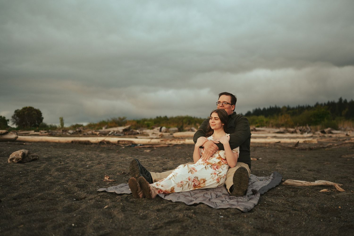 A couple relaxes together on a blanket at a moody beach with driftwood and dramatic cloudy skies.