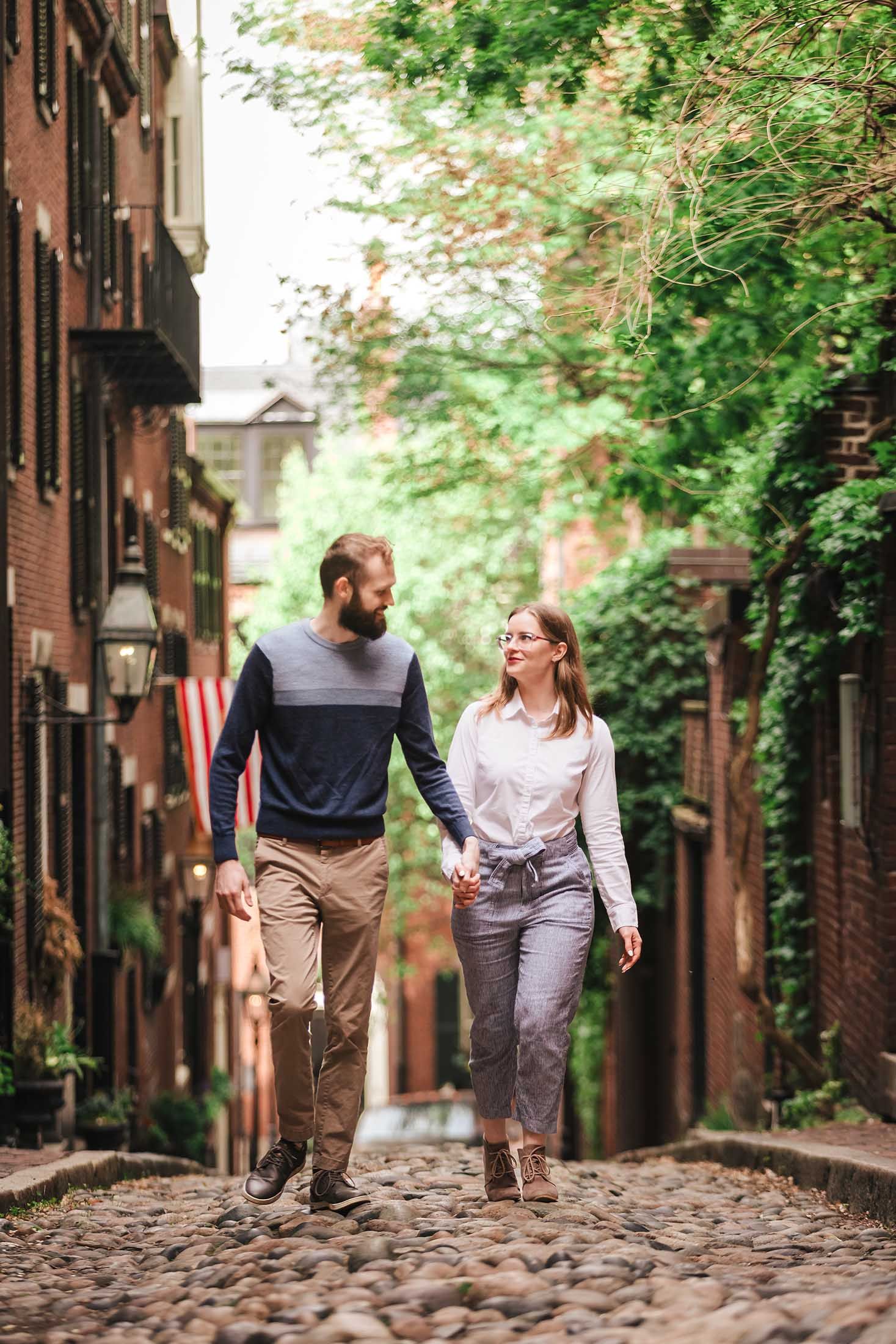A couple strolls side by side along the charming cobblestones of Acorn Street, smiling at one another amid Boston’s iconic brick facades