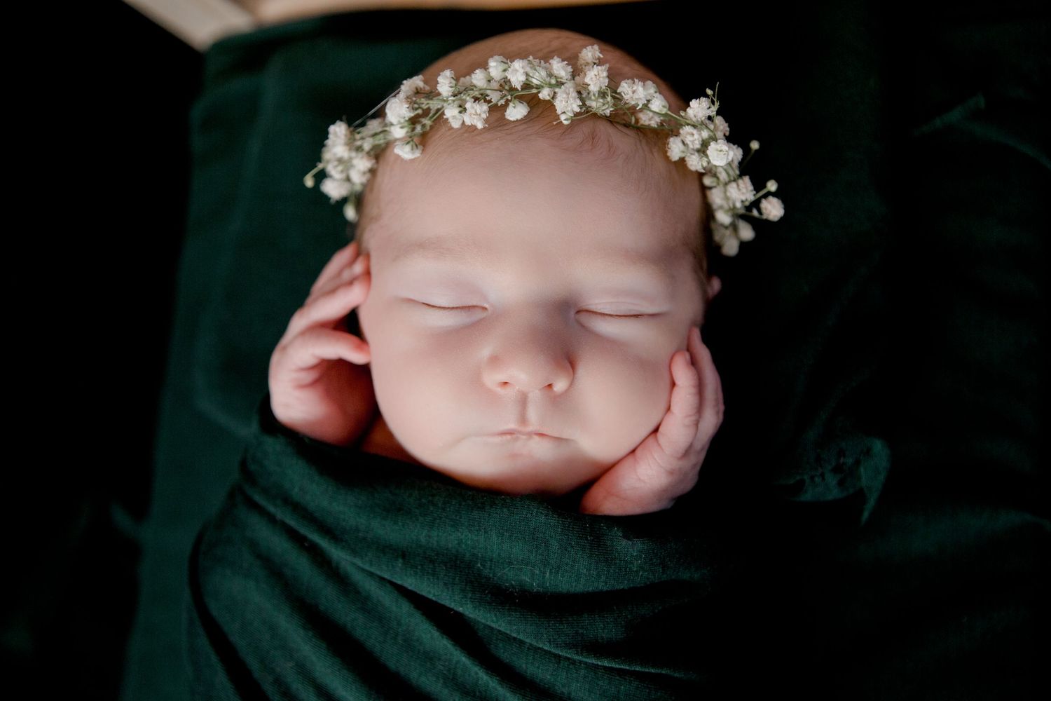 Sleeping newborn wearing delicate flower crown on dark green backdrop.