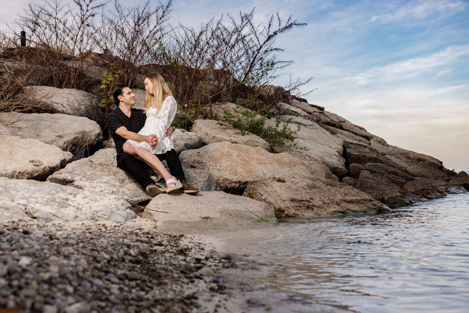 Person sits on rocky coastal outcrop reading a book near the water's edge at sunset.