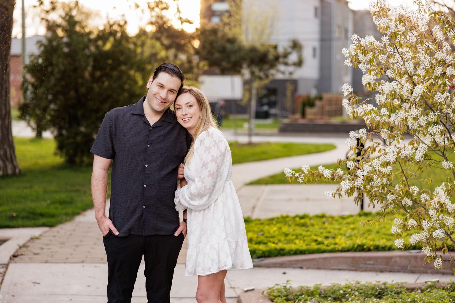 A couple stands together on a sidewalk in a garden setting with blooming white flowers in springtime.