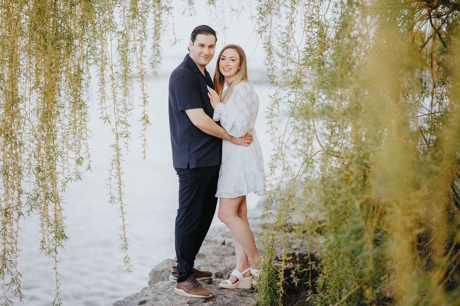 A couple embraces under a romantic canopy of drooping willow tree branches at sunset.