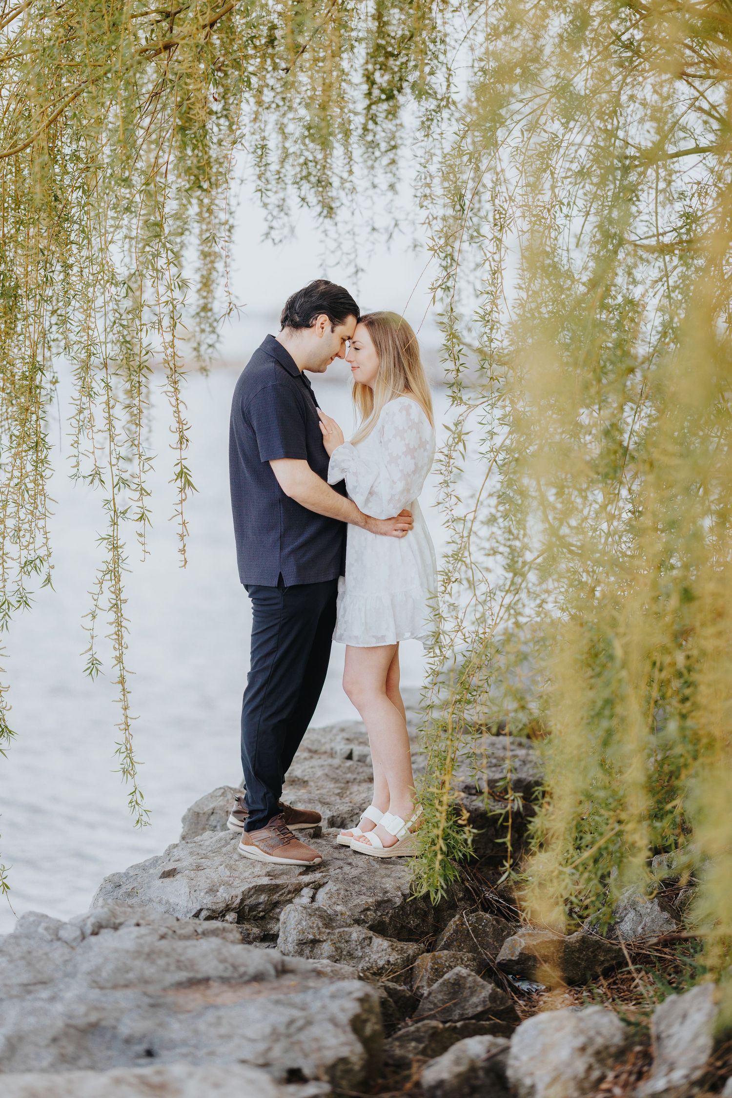 Romantic couple sharing intimate moment together on rocky shoreline under cascading willow branches at sunset.
