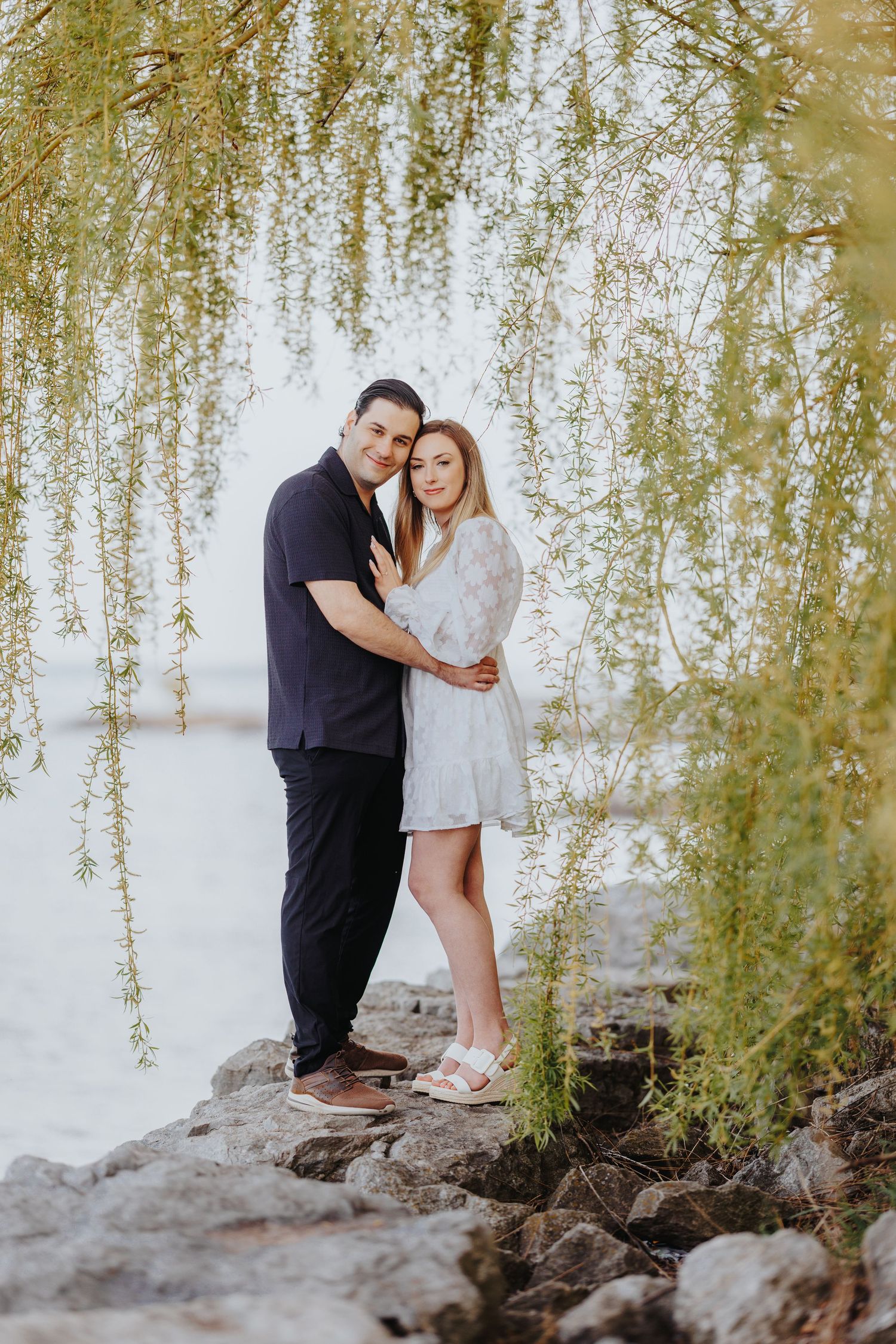 A romantic couple embraces under cascading willow branches while standing on rocky terrain near a white wall.