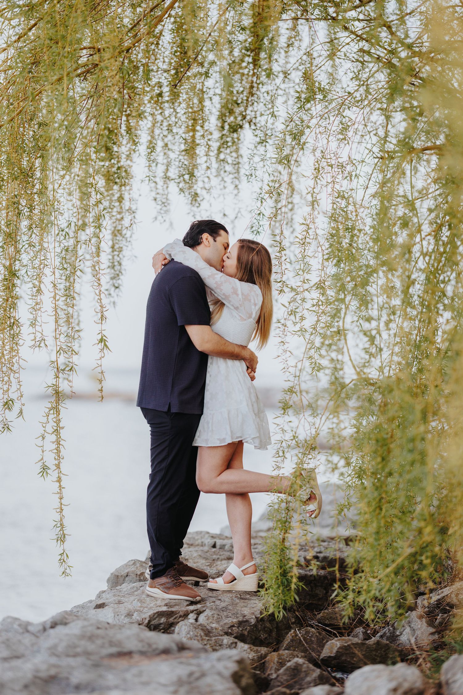 A tender moment captured under weeping willow trees as a couple shares a kiss while standing on rocks.