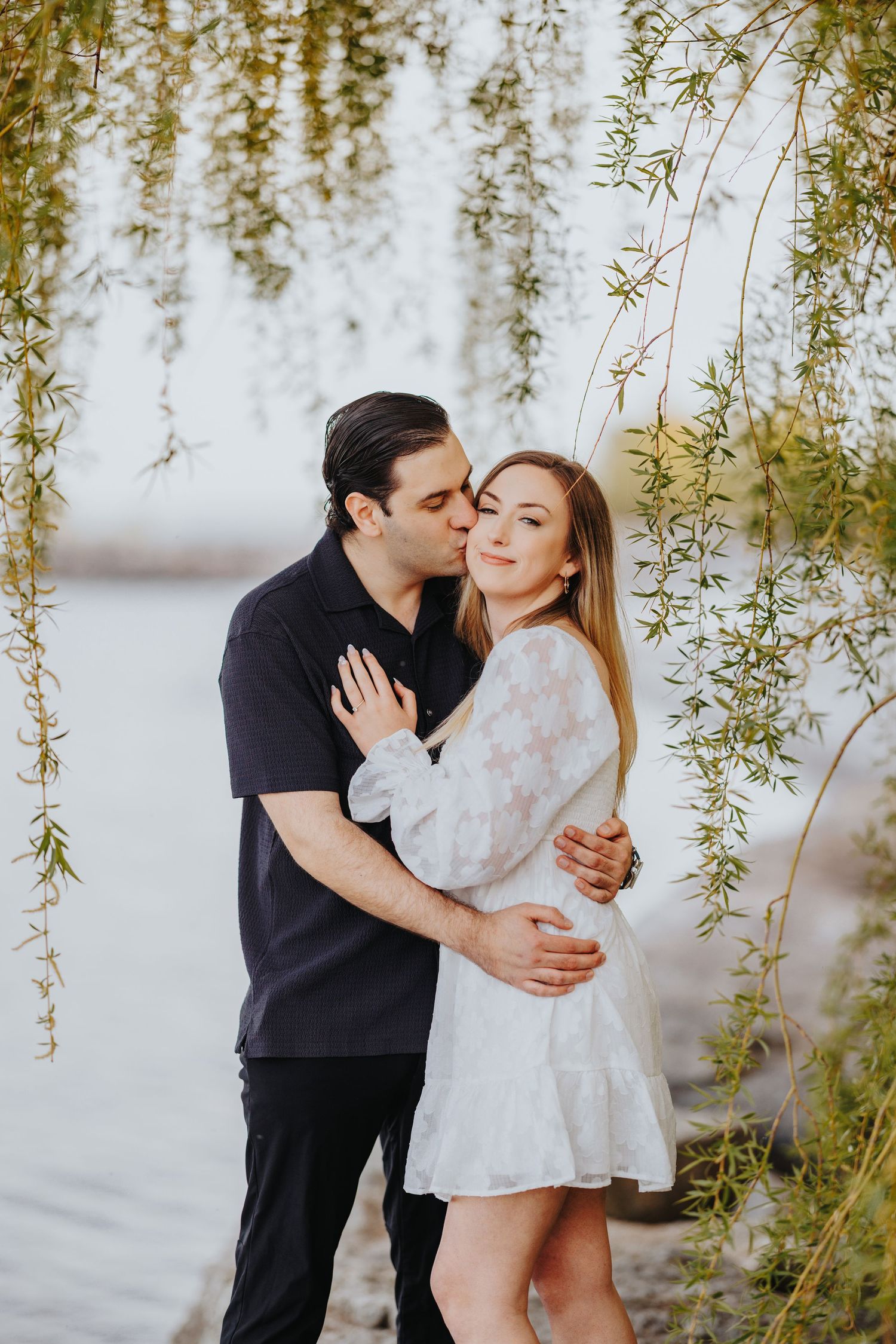 Romantic couple embracing under drooping willow tree branches near a lake.