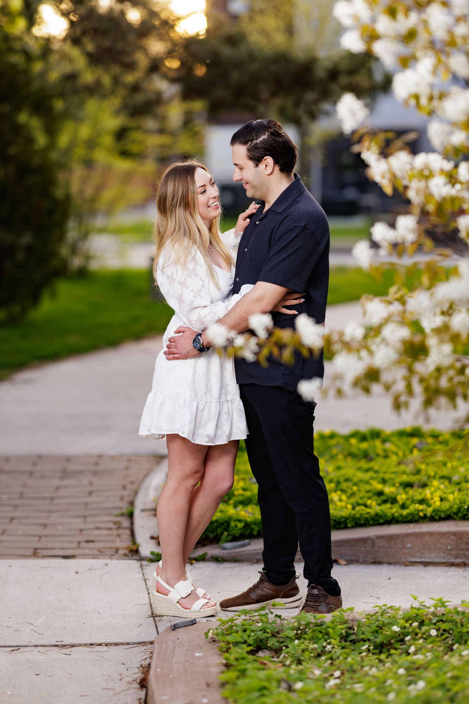 A romantic couple embracing on a garden path during sunset with white spring blossoms in the foreground.
