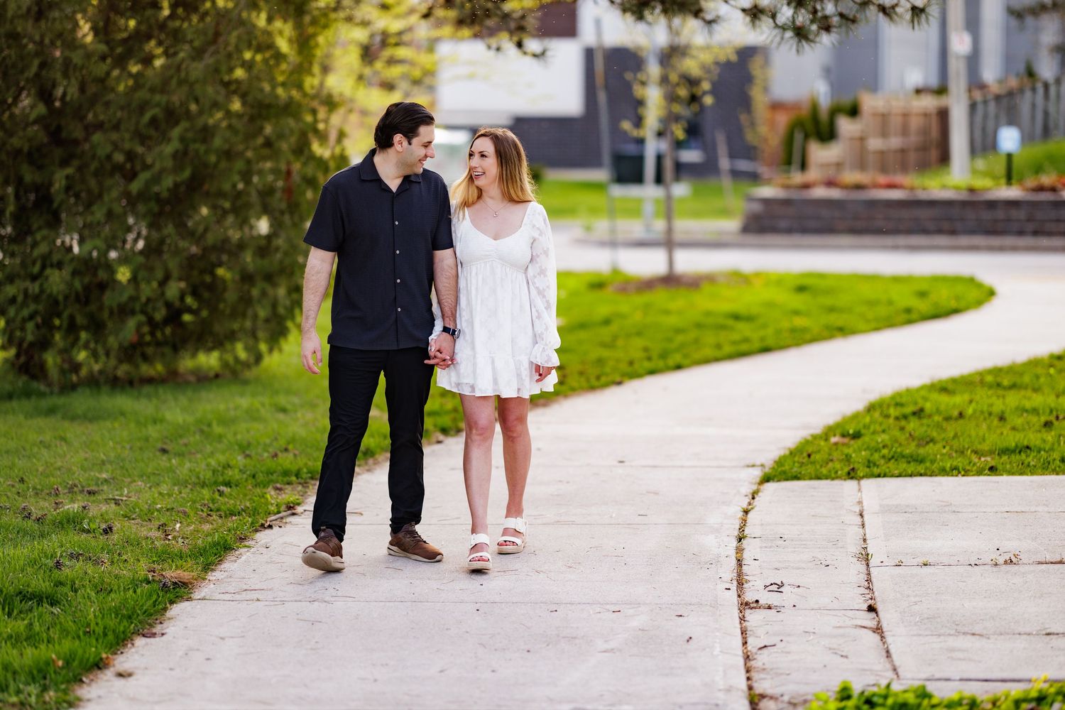 Two people walk together on a curved concrete sidewalk through a landscaped park area on a sunny day.