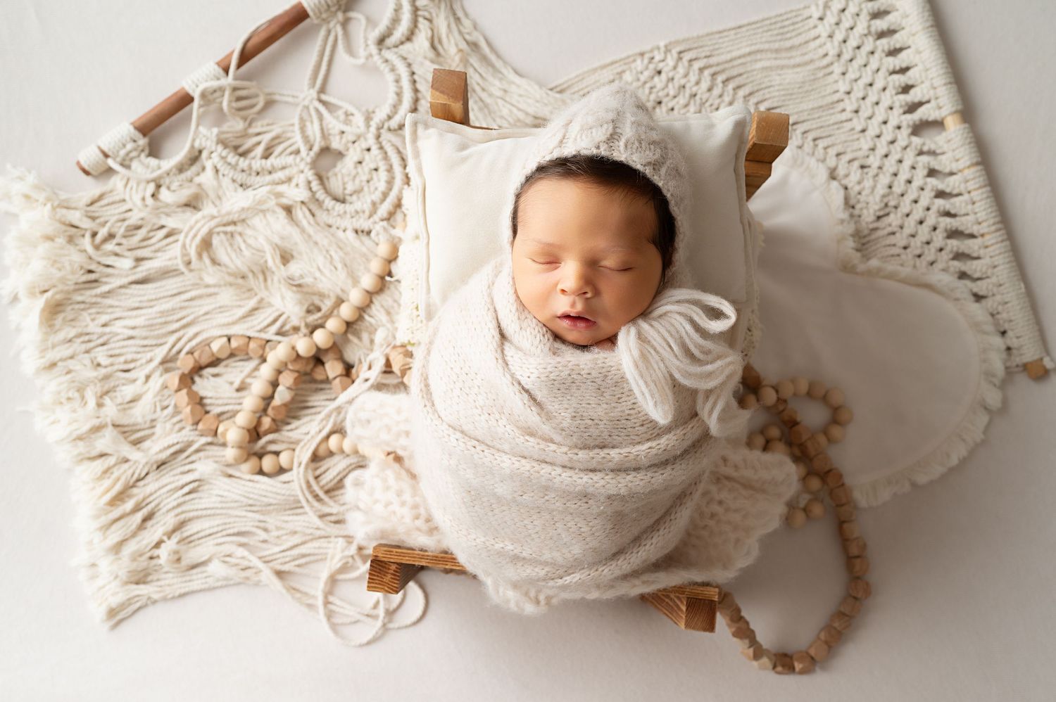 Newborn baby wrapped in white swaddle on macrame blanket with decorative elements.
