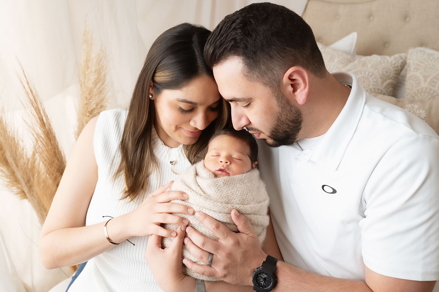 Parents cradling their newborn baby surrounded by soft pampas grass in a natural light setting.