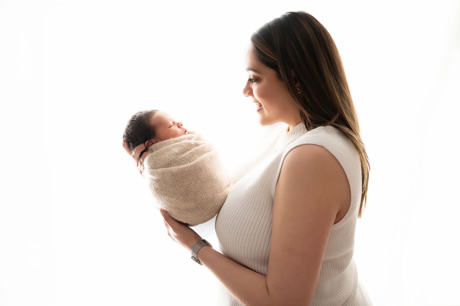 A mother in a white dress gazes lovingly at her newborn baby wrapped in a beige swaddle against a light background.