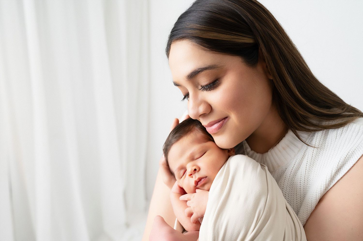 A tender moment of a person in white embracing a smiling baby against a light background.