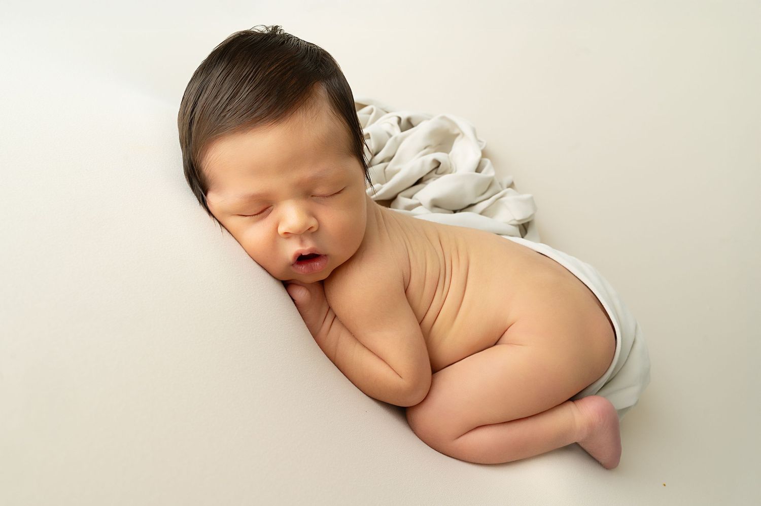 Sleeping newborn posed on side with hands tucked under face on white backdrop.