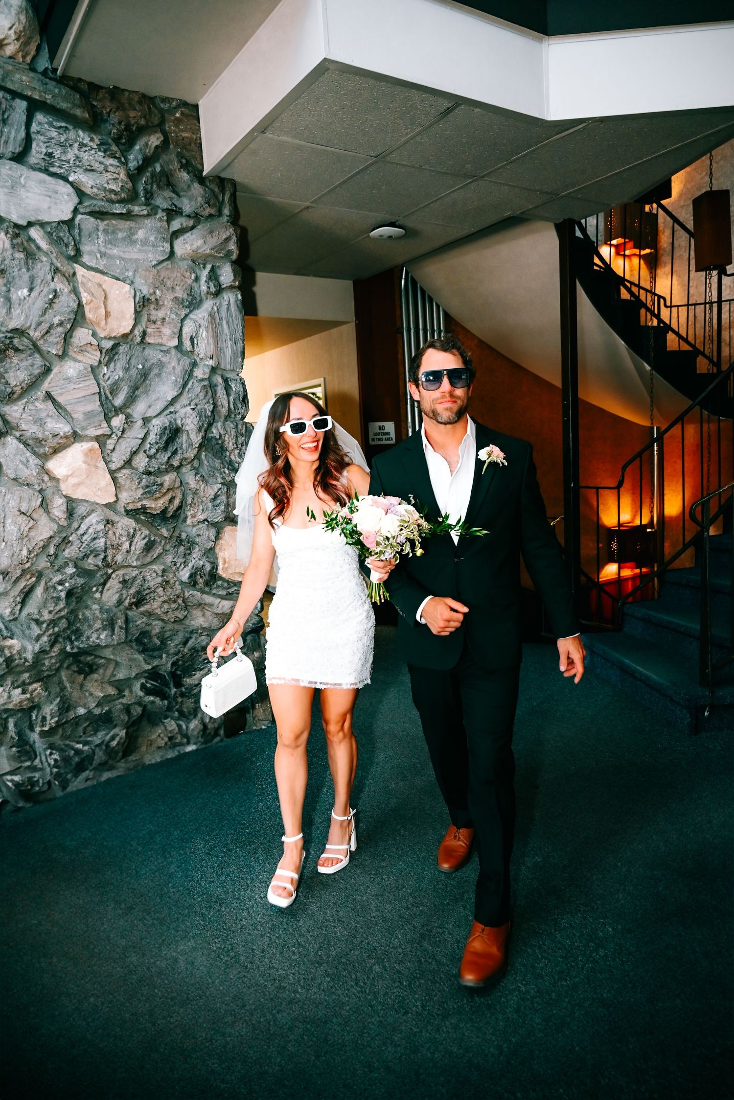 A couple in formal attire walk through a hallway with stone walls and dark carpeting.