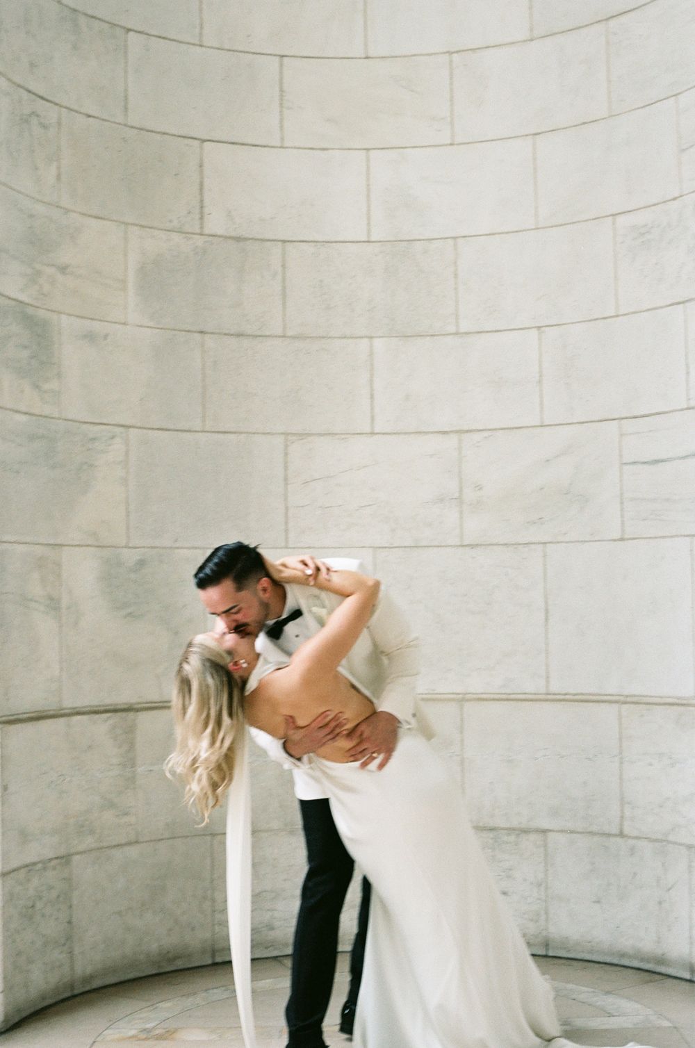 A romantic couple in wedding attire share an intimate dance pose under a curved stone wall. paris film wedding photographer