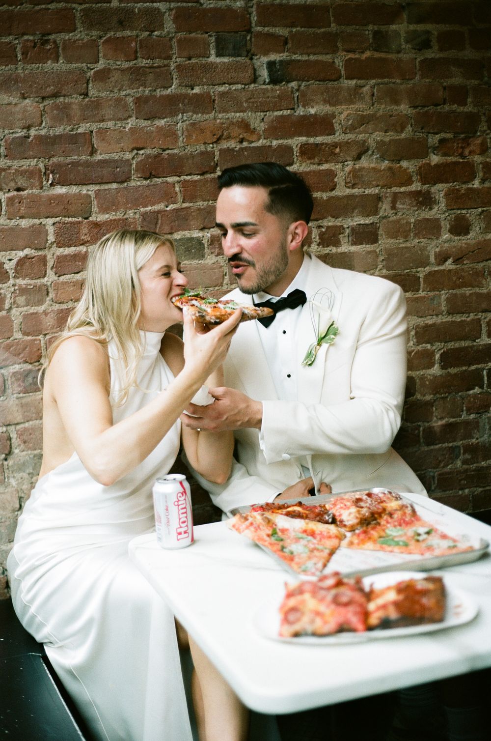 A couple in wedding attire sharing pizza slices at a casual reception against an exposed brick wall. paris wedding eating pizza, film