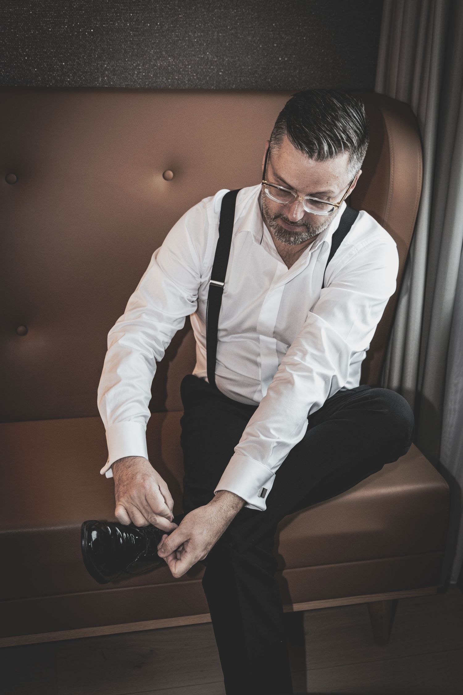 Groom in white shirt and suspenders putting on dress shoes while seated in leather chair.