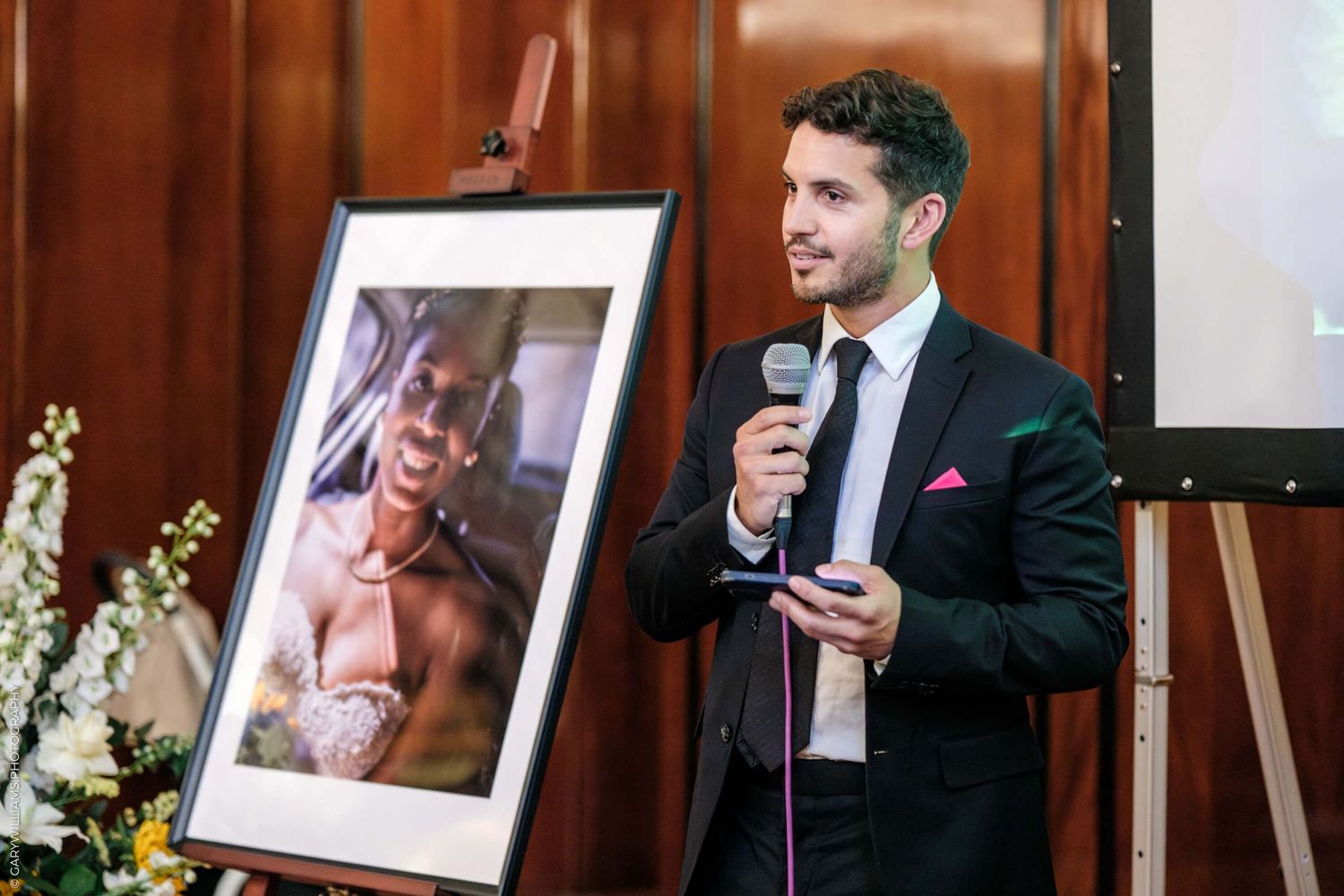 Speaker stands at podium beside framed artwork during formal event with wooden paneled backdrop and floral arrangements.