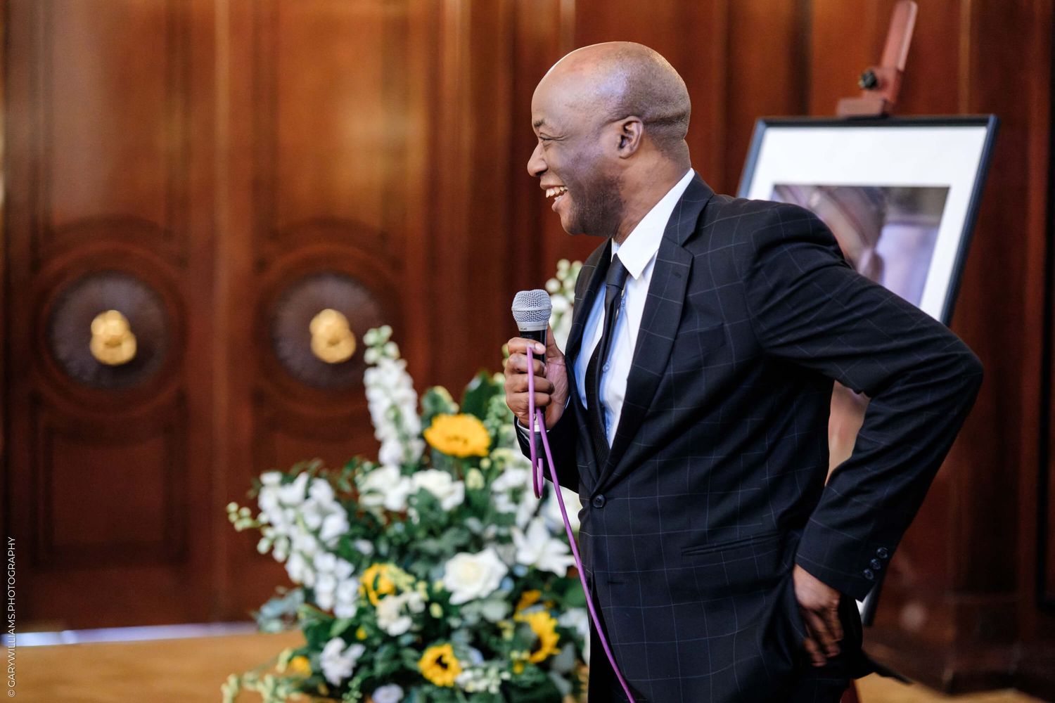 An entertainer in a black suit performs on stage with a microphone next to floral arrangements in an elegant venue.