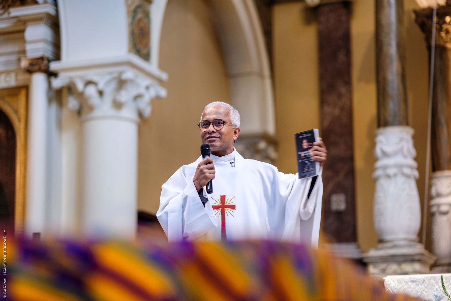 A person in white clerical vestments holds a microphone and mobile phone at a church pulpit with ornate columns visible.