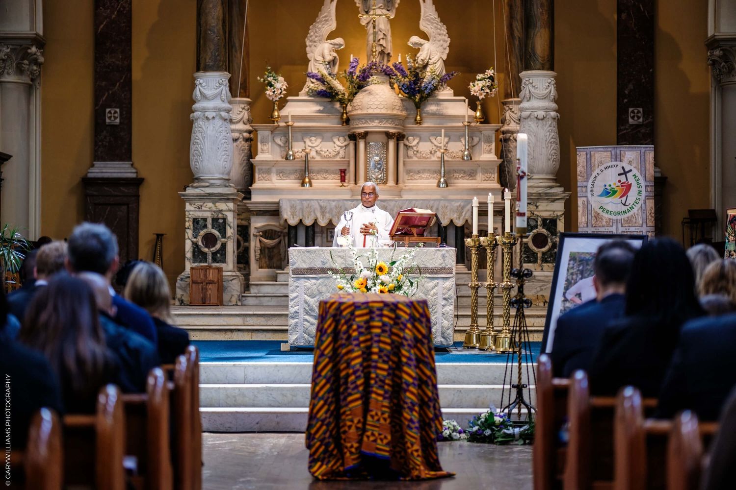 Interior view of a Catholic church during a mass service with ornate altar, candles, and congregation in wooden pews.