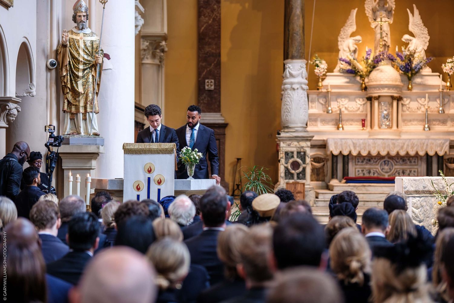 A speaker addresses a large audience from a podium inside an ornate church with golden decorative details and religious statues.