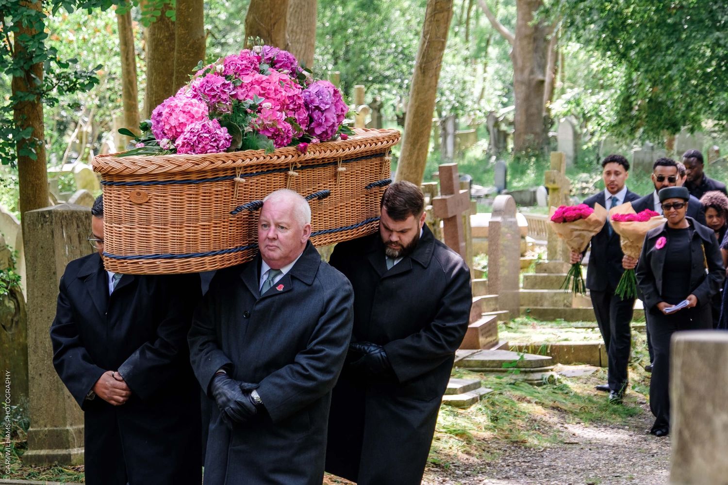 Pallbearers carrying a pink flower-adorned wicker casket through a wooded cemetery path during a funeral procession.