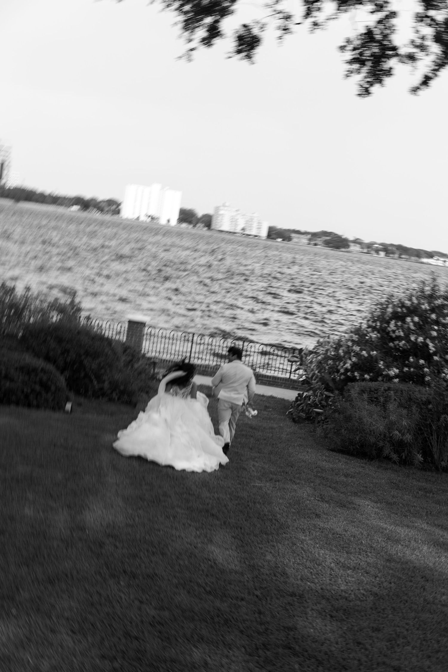A bride in a flowing white gown holds hands with her groom as they walk toward a waterfront in black and white.