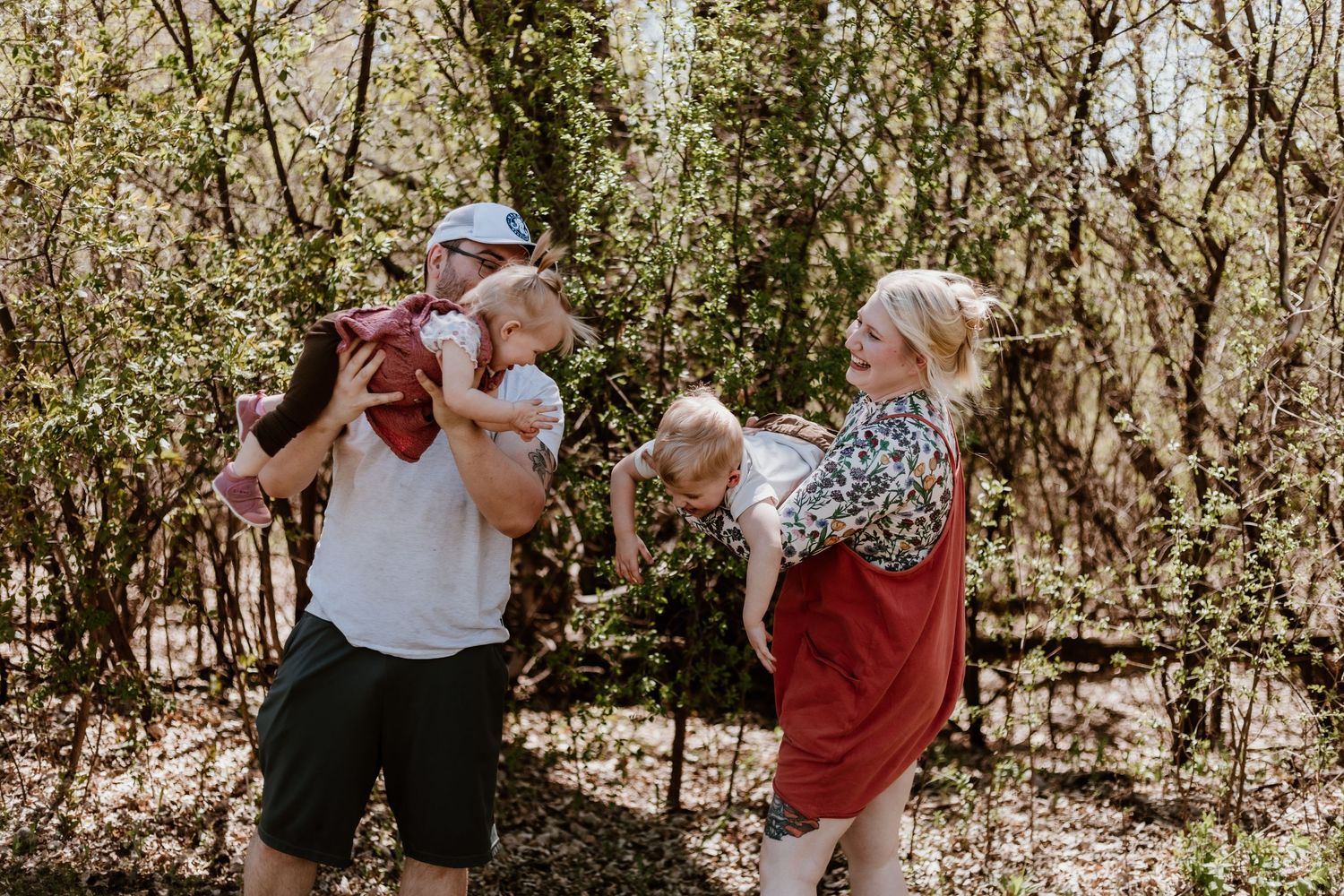 Family walking through a wooded hiking trail on a sunny day with young children on their shoulders.