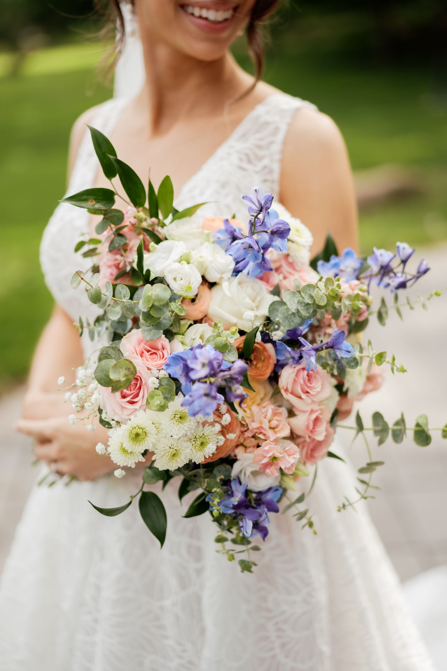 A delicate bridal bouquet featuring pink roses, white flowers, purple delphinium and eucalyptus against a lace wedding dress.
