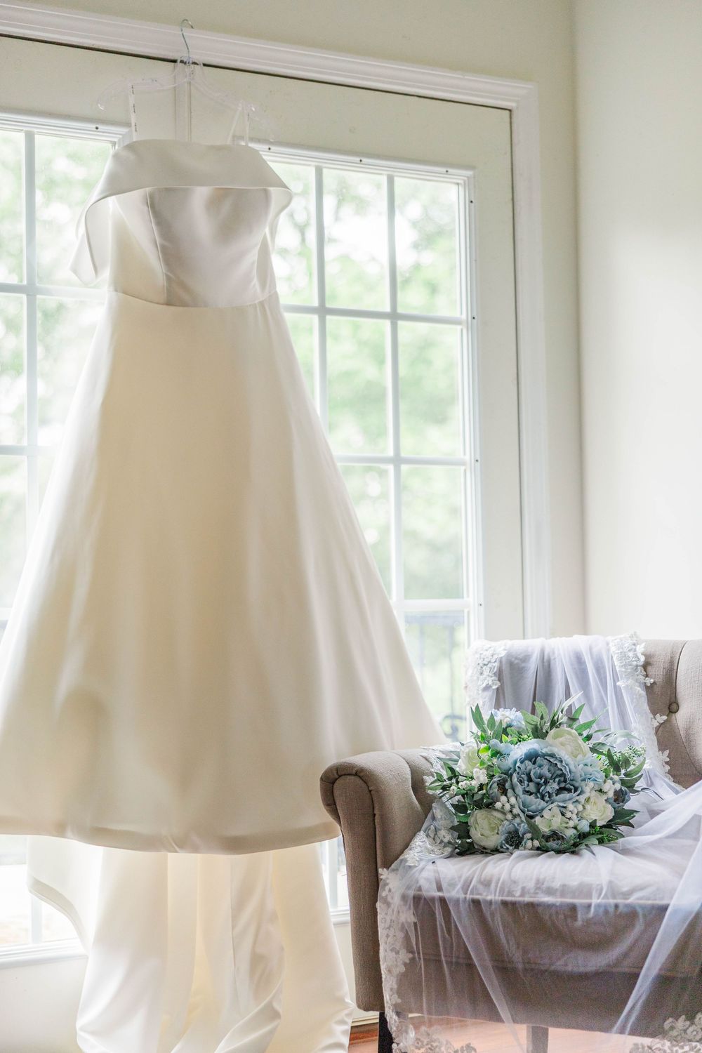 Elegant white wedding gown hanging in front of bright windows with a blue bouquet on chair.