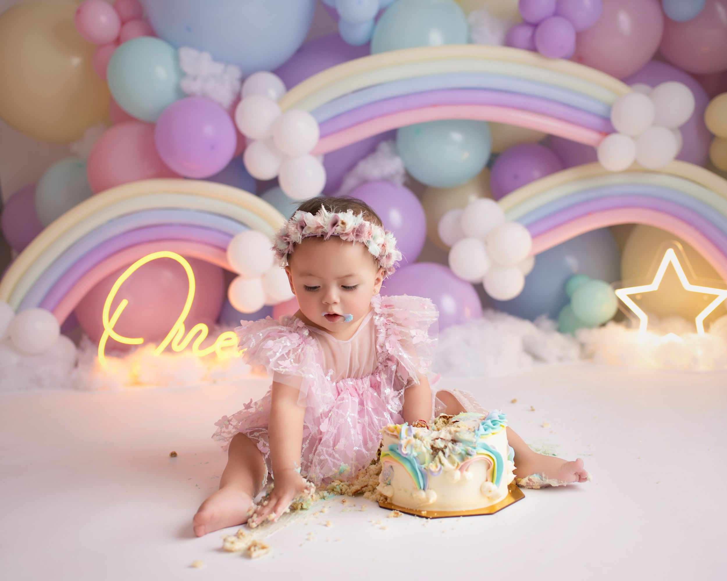 A cake smash photo session with pastel balloons, neon signs, and rainbow decorations in dreamy pink tones.