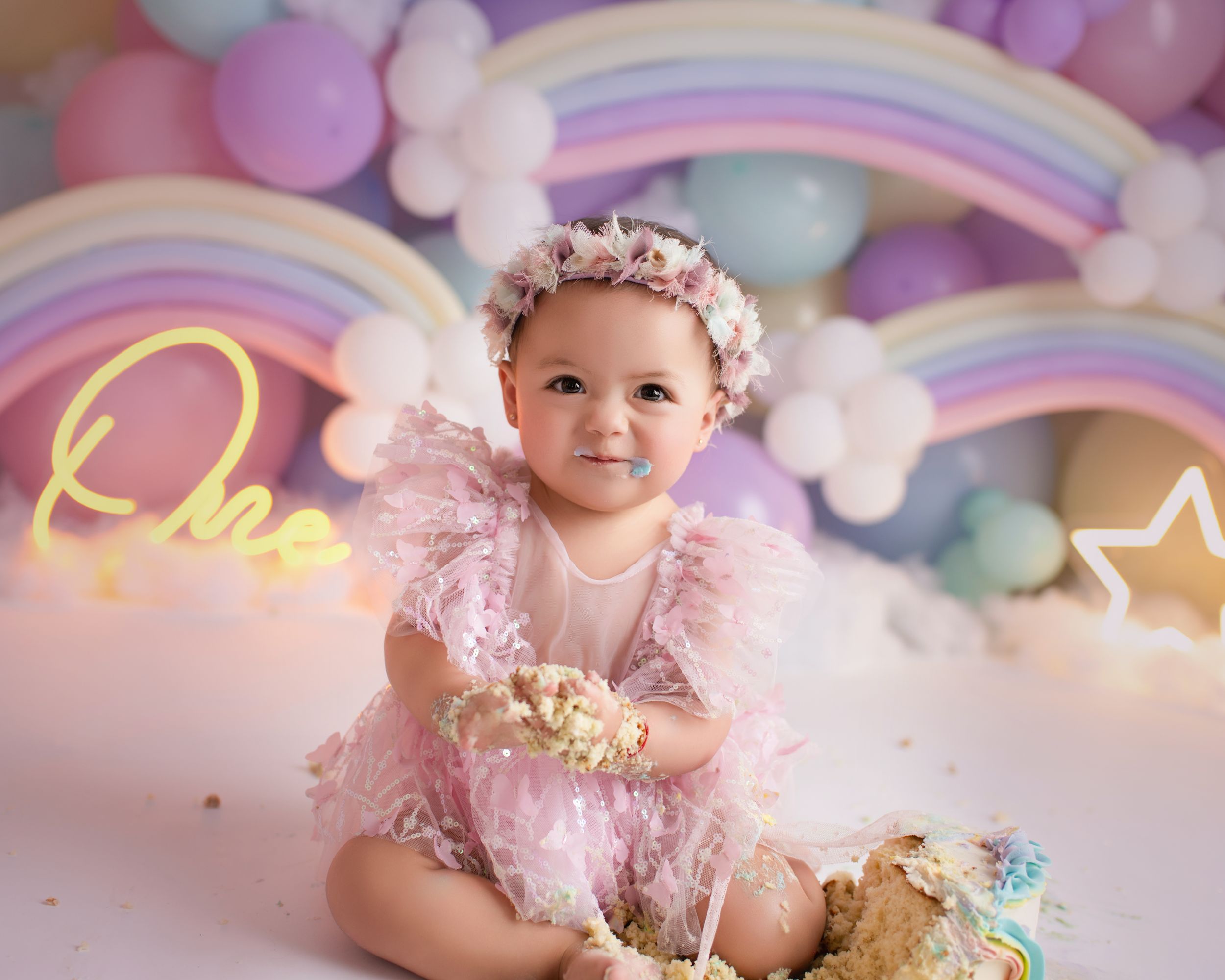 Whimsical first birthday photoshoot featuring pastel decorations, neon lights and a rainbow backdrop.