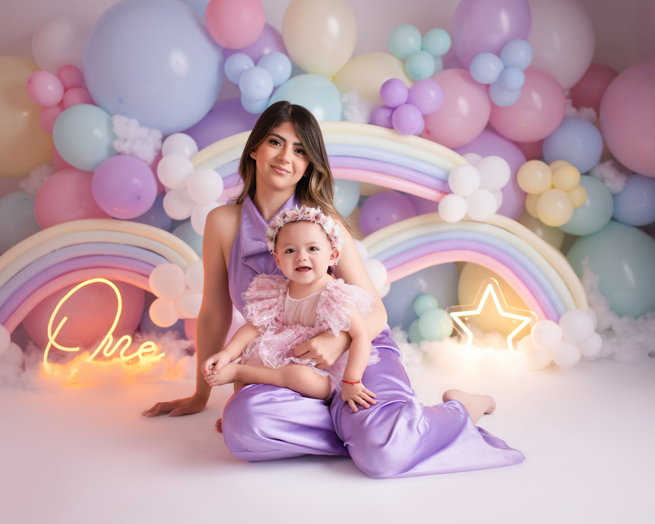 Whimsical cake smash setup with pastel balloons, neon rainbow sign, and purple decorations.
