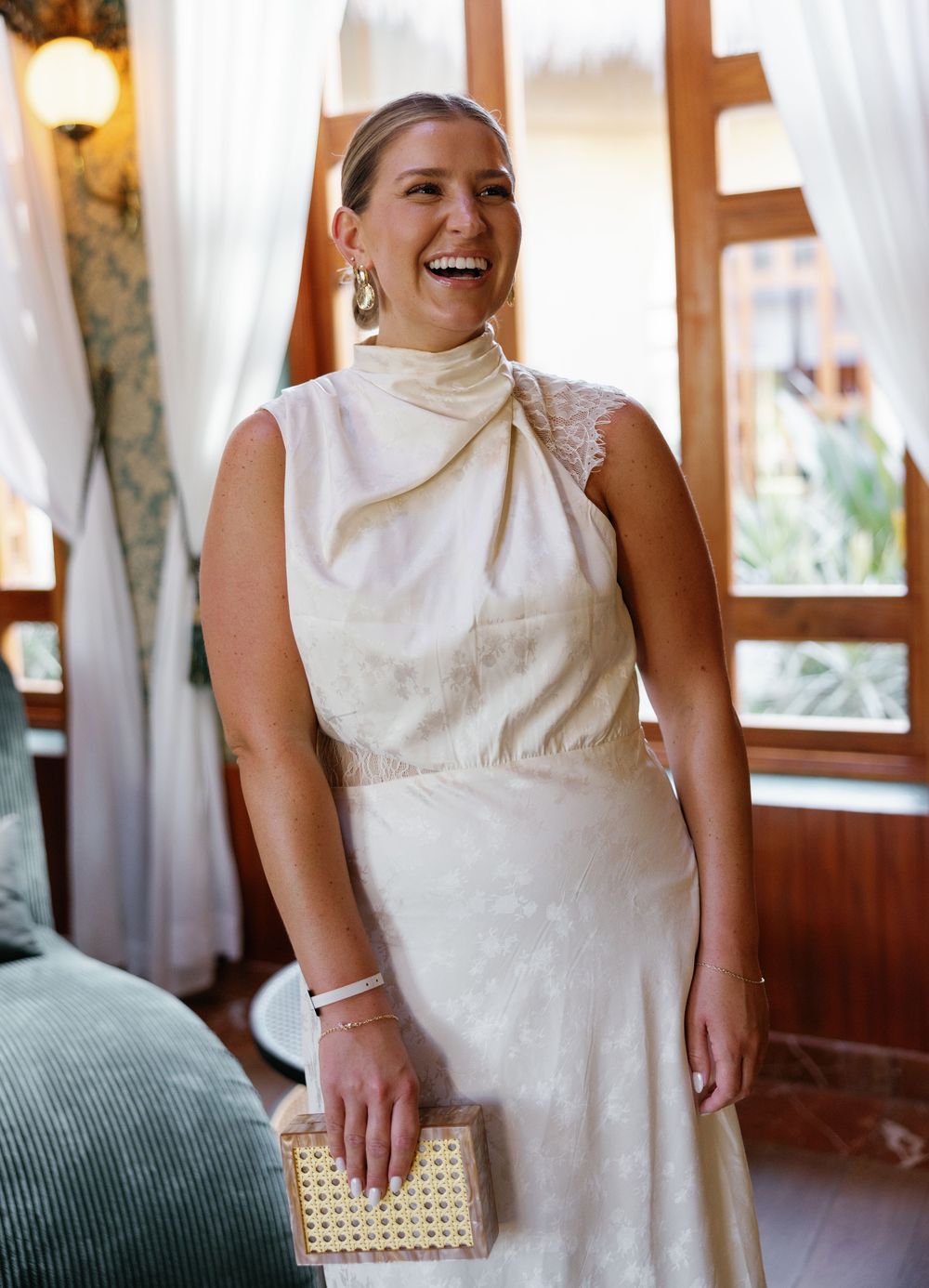 Elegant guest wearing a cream-colored high-neck sleeveless dress with a woven clutch at an indoor venue.