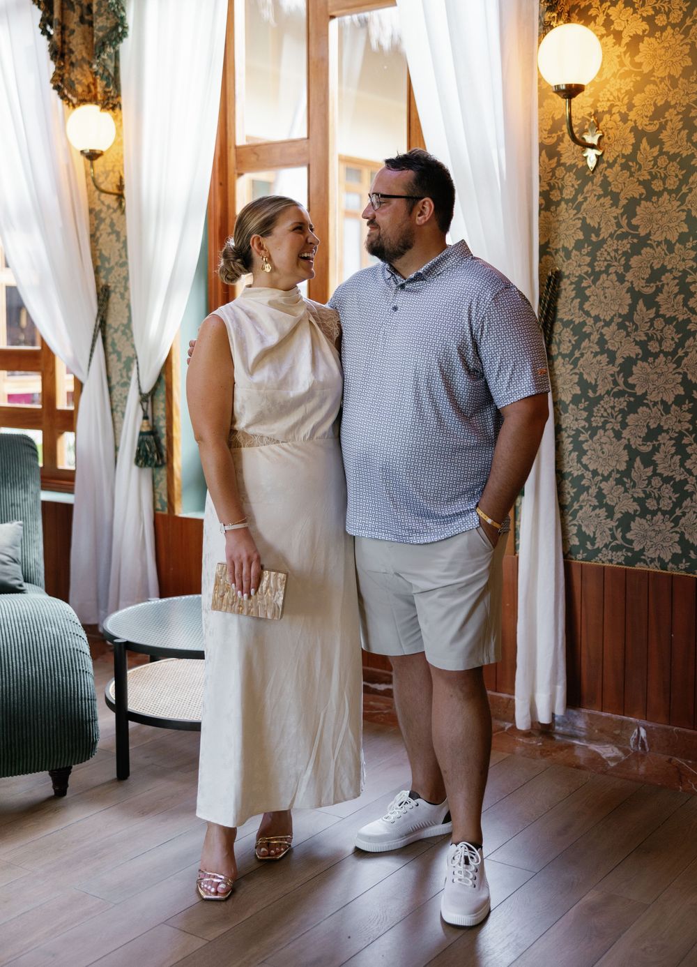 A couple in elegant attire standing together in an elegant room with sheer curtains and vintage wallpaper.