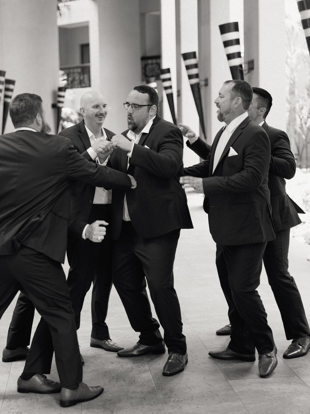 Black and white photo of groomsmen in suits sharing a candid laughing moment at a wedding venue.